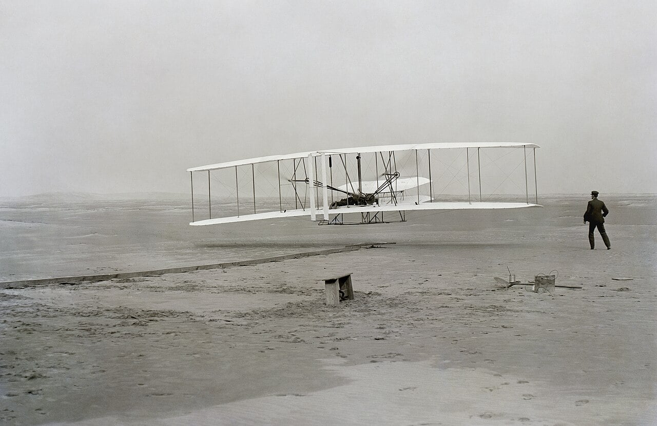 The Wright Flyer lifts off at Kitty Hawk, December 17, 1903 — Orville at the controls, Wilbur running alongside. The 12-second flight that changed human history.