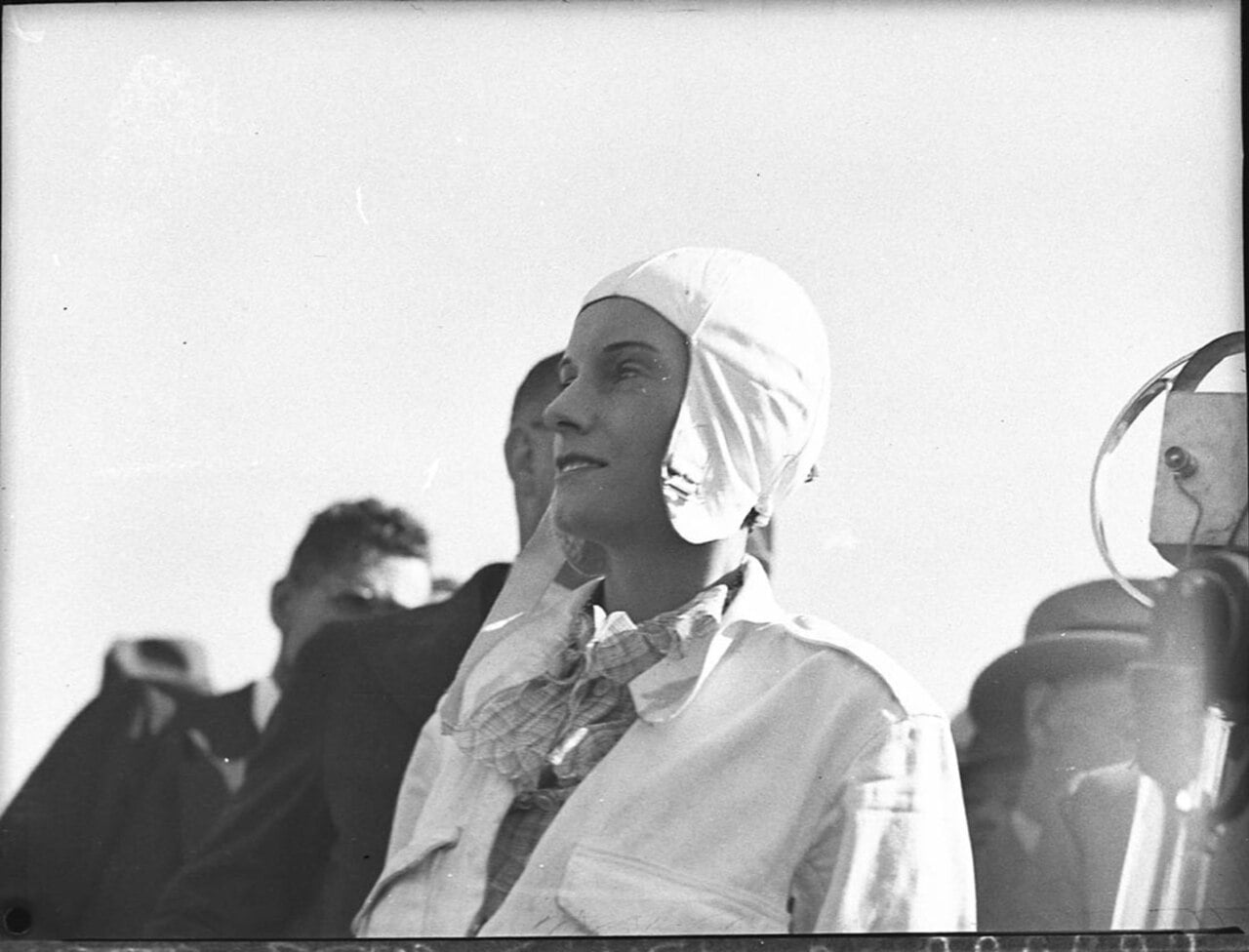 Jean Batten in flying helmet speaking at a microphone on arrival