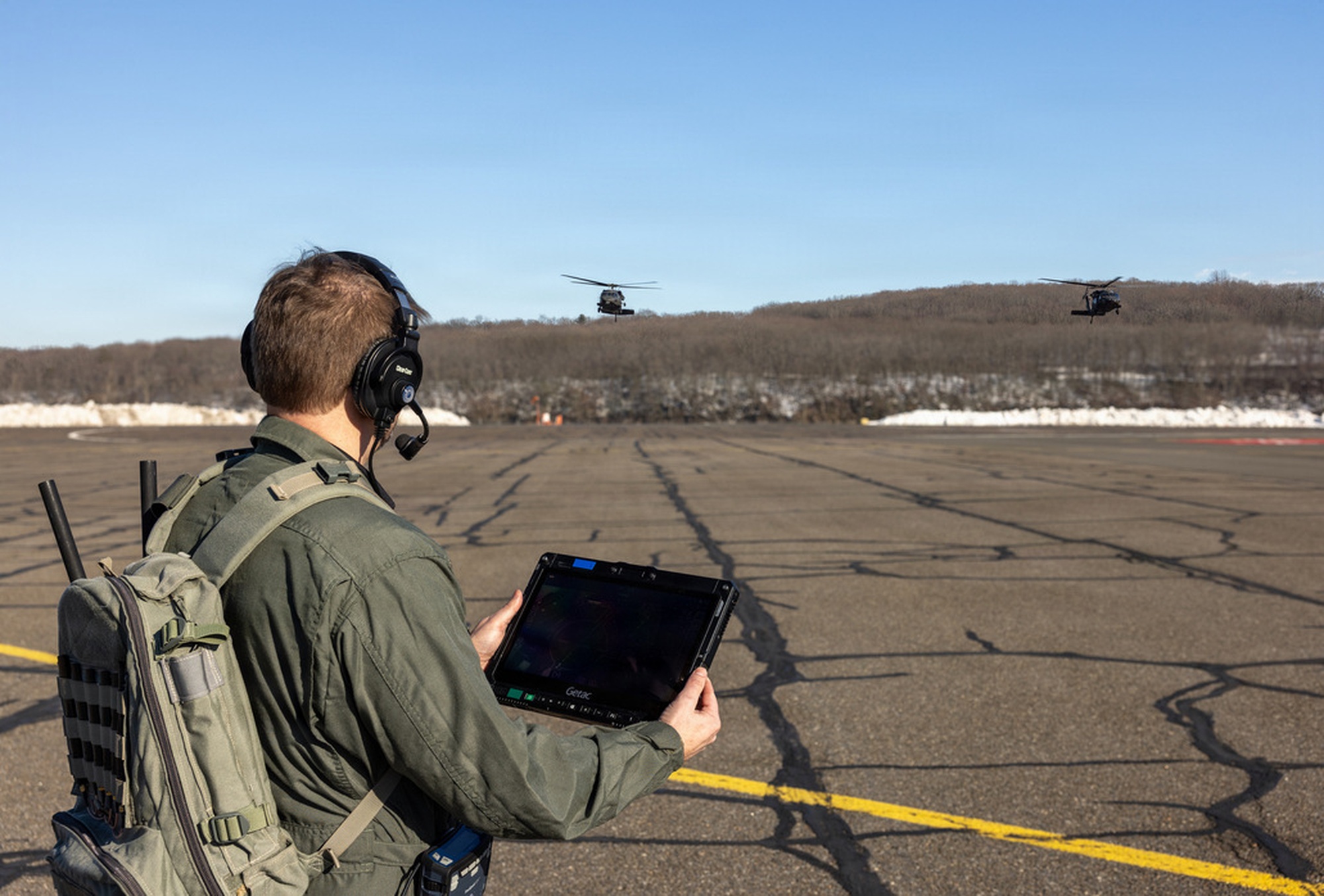 An operator on a flight line monitors two Black Hawk helicopters using a hand-held tablet