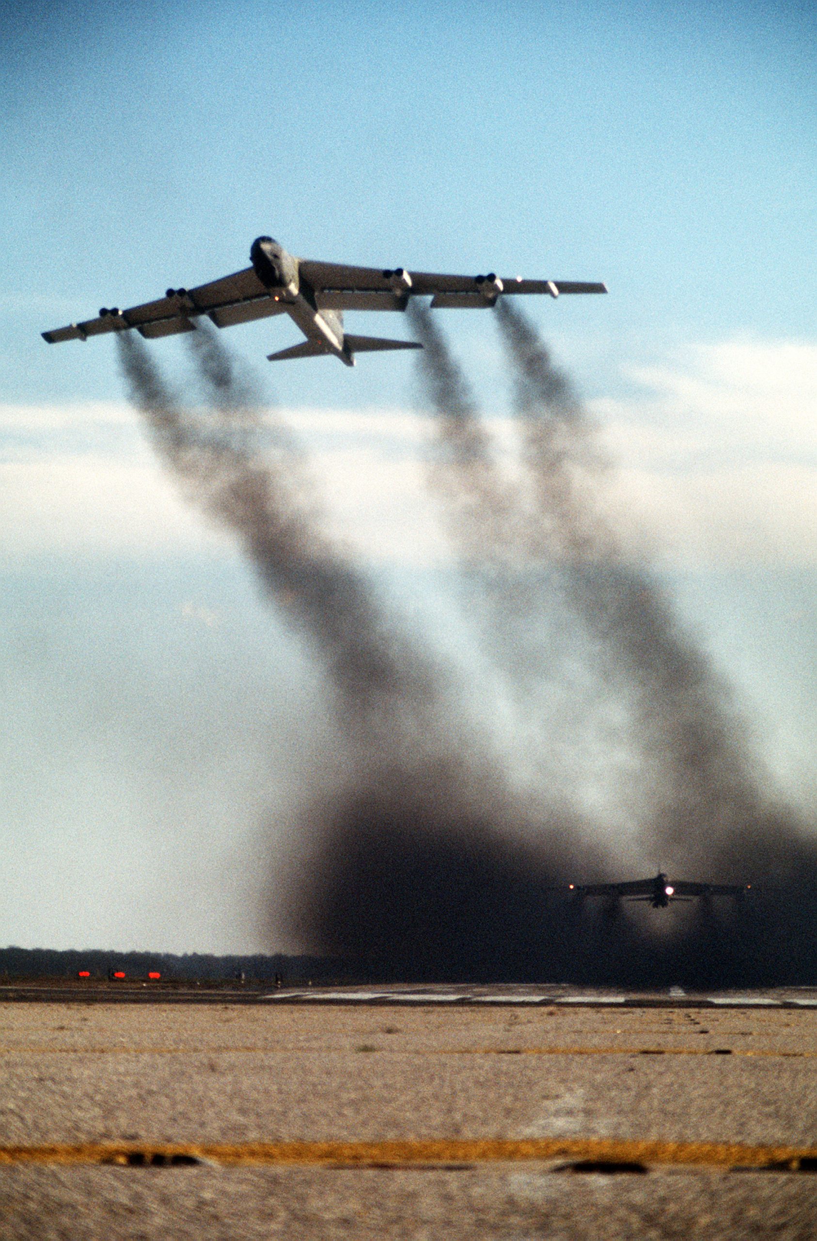 A B-52G climbing away trailing heavy black smoke while a second B-52 rolls in the smoke behind it