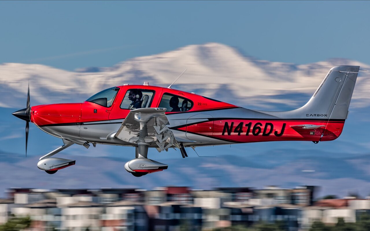 A Cirrus SR22 single-engine aircraft in flight against a mountain backdrop