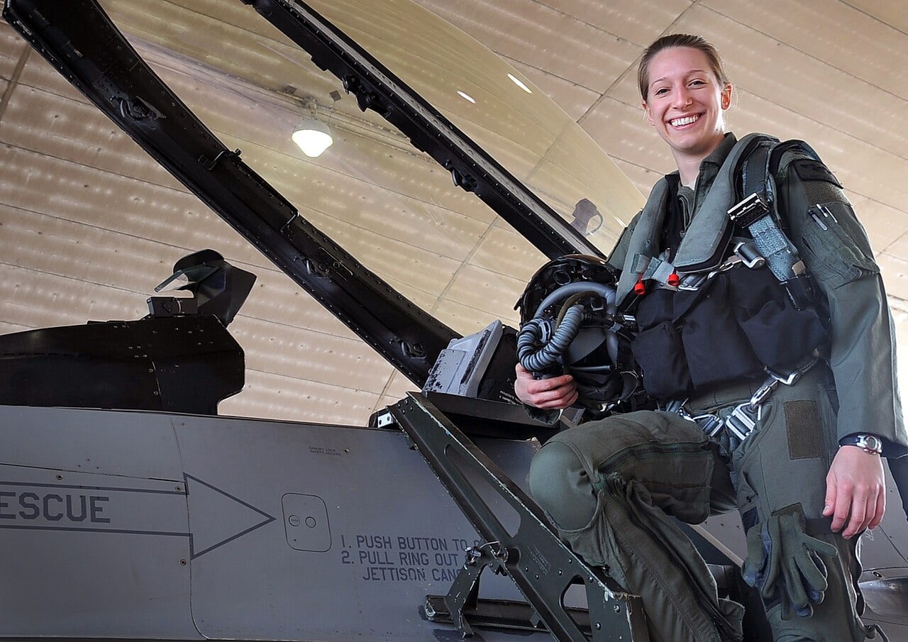 A US Air Force pilot climbing into an F-16 cockpit wearing a parachute harness with leg straps visible