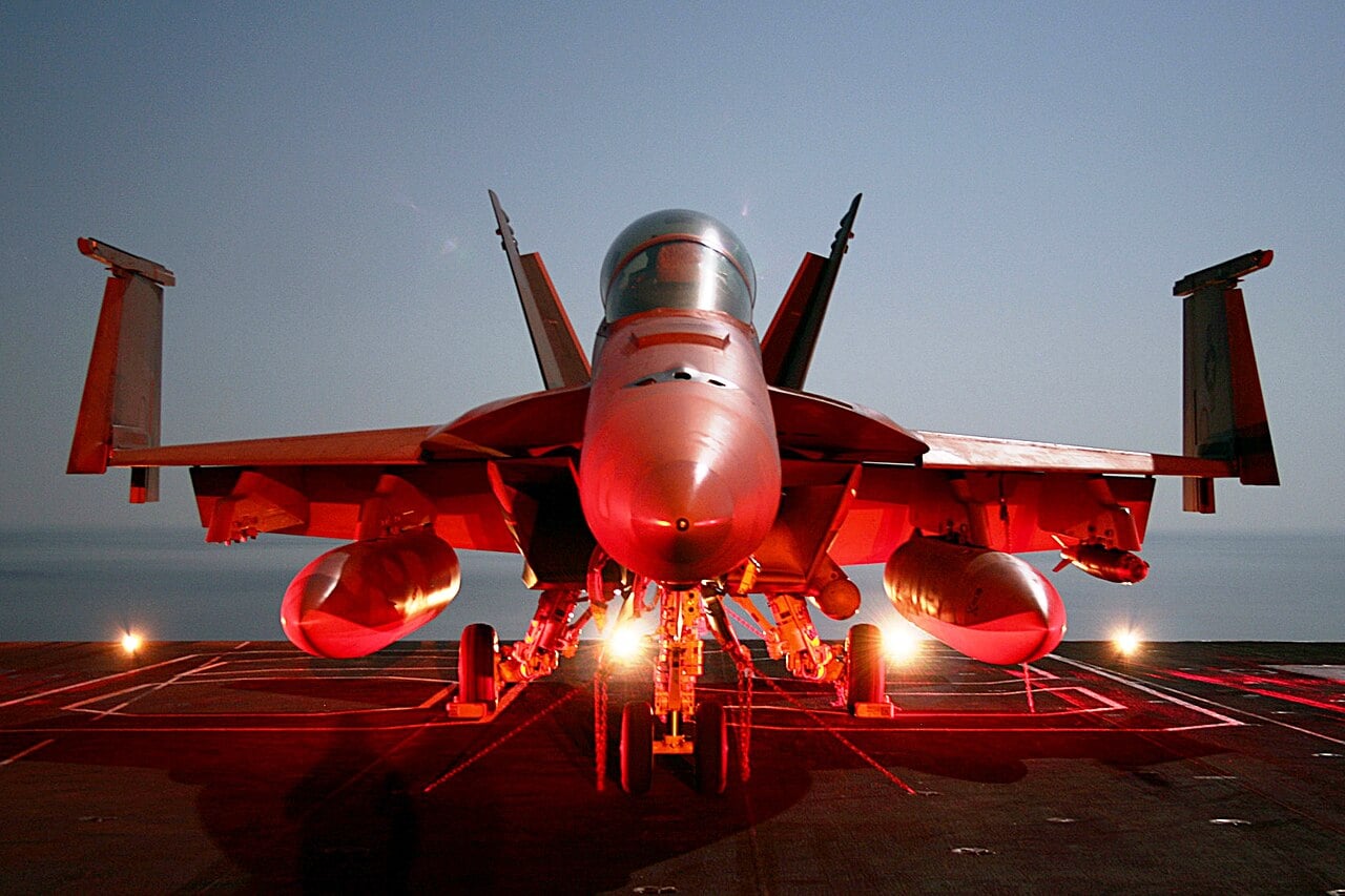 An F/A-18 Super Hornet chained to the flight deck with its wingtips folded upward under red deck lighting