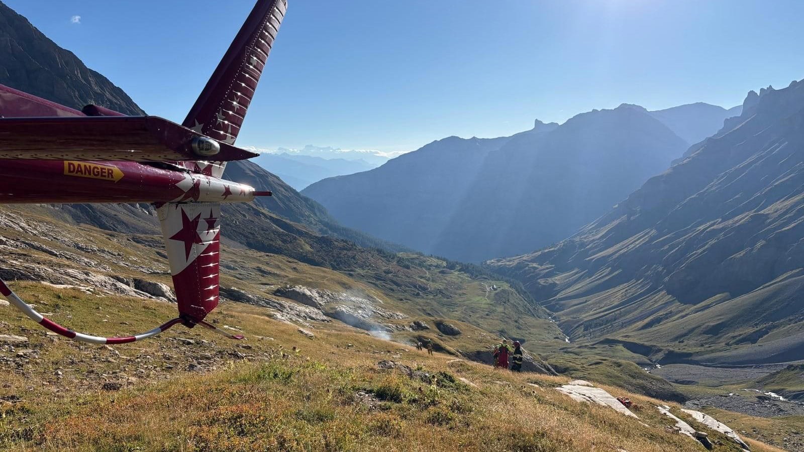 A rescue helicopter tail in the foreground with the Fluhalp basin above Leukerbad and responders on the slope
