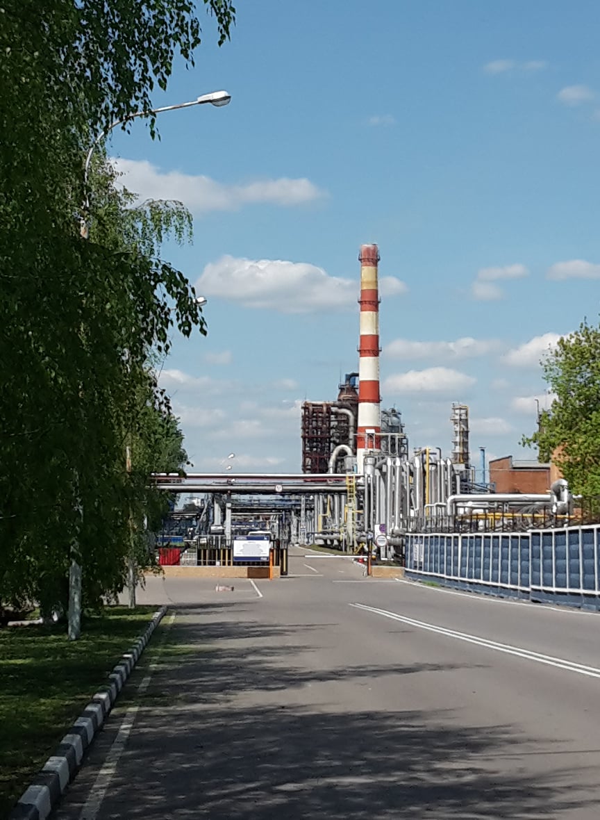 The Gazprom Neft Moscow Oil Refinery seen from the approach road, with its stack visible above the treeline