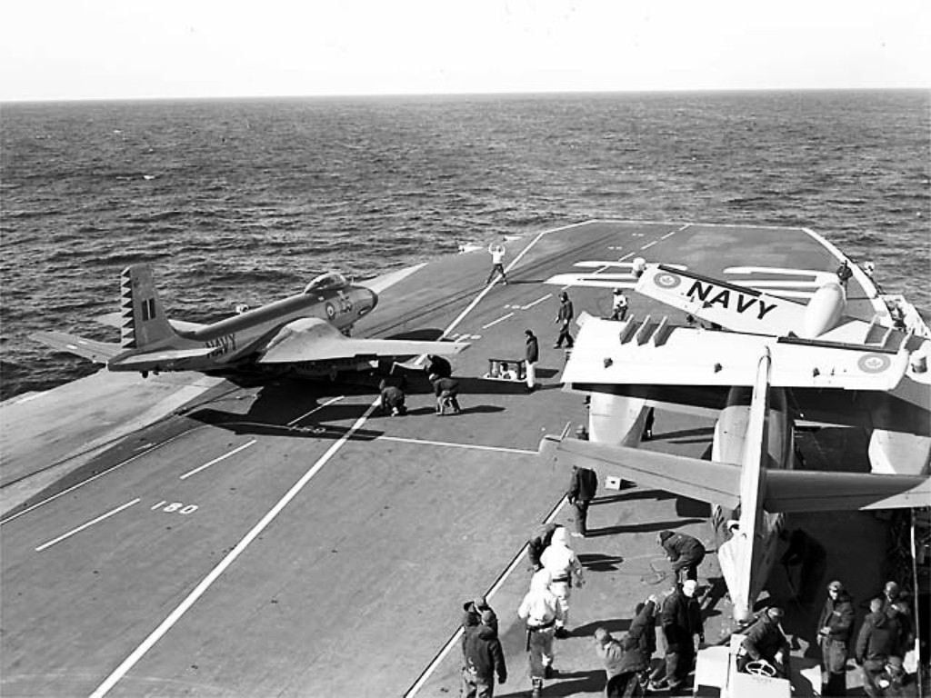 A Banshee sits on the catapult of HMCS Bonaventure with a Tracker parked forward, 1957
