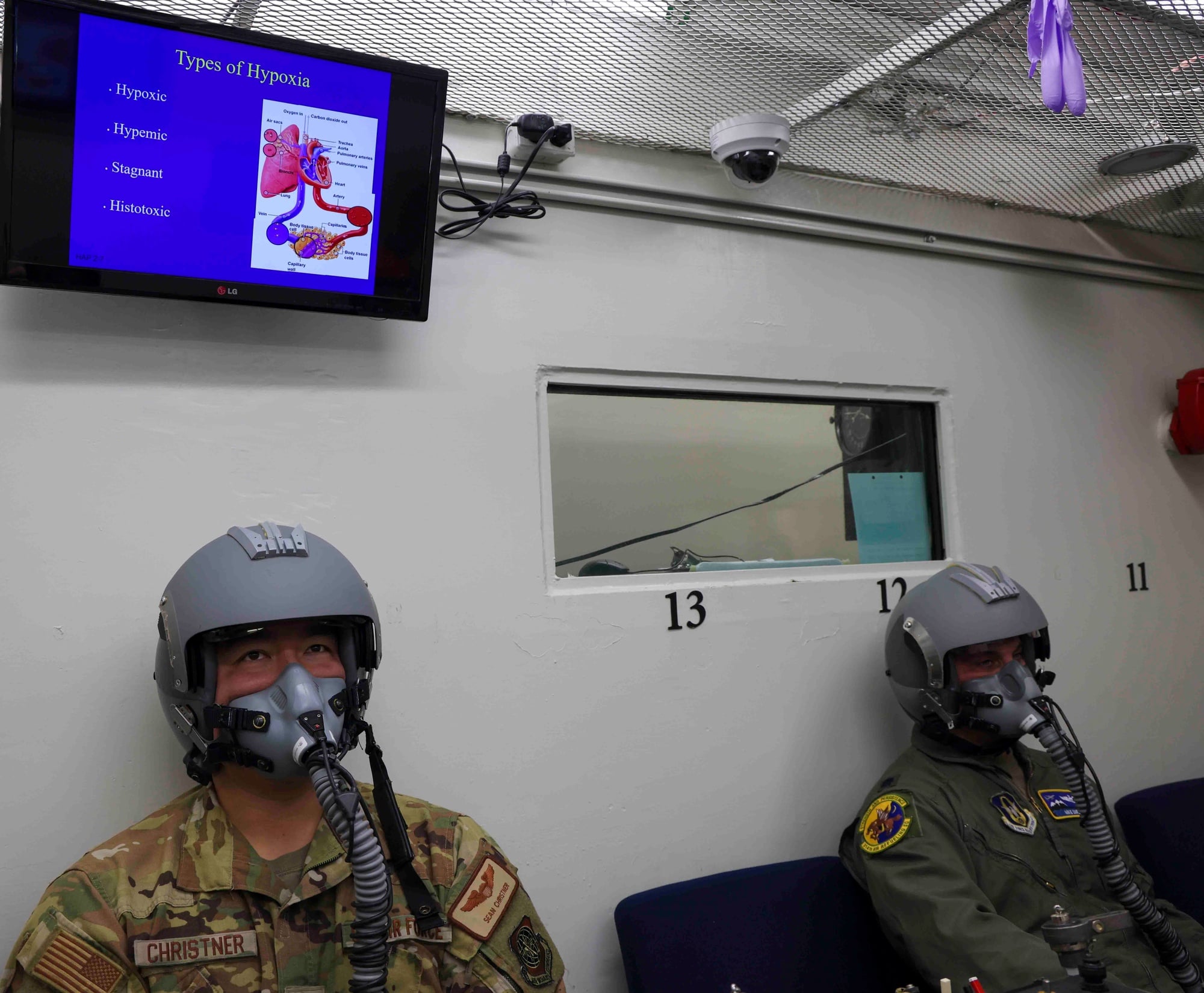 Two aircrew students on oxygen masks beside a Types of Hypoxia briefing screen in an altitude chamber