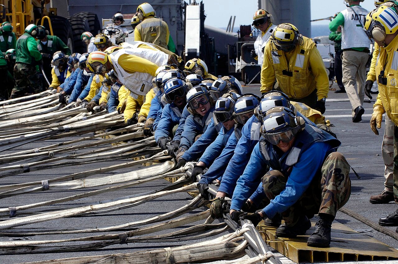 Blue, yellow, green and white shirted US Navy flight deck crew erecting a crash barricade