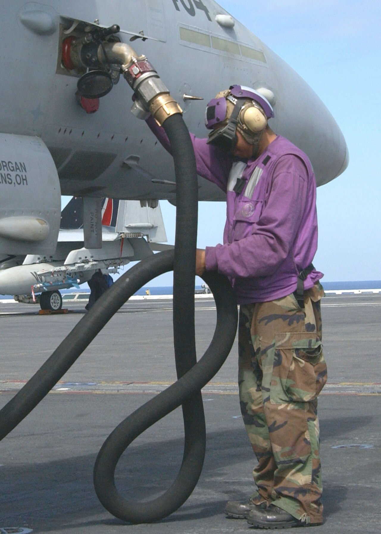 A US Navy aviation fuel handler in a purple jersey refuelling an aircraft on a carrier flight deck