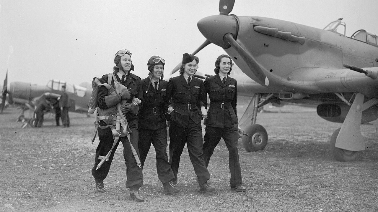 Four Air Transport Auxiliary women pilots walking arm in arm past a Hawker Hurricane in March 1943