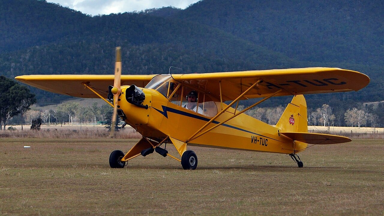 A yellow Piper J-3 Cub taildragger on a grass airfield with hills behind