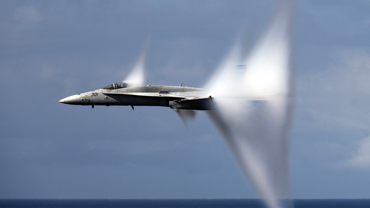 F/A-18C Hornet with a small vapor cone at the canopy and a large one behind the wing, flying low over the Pacific