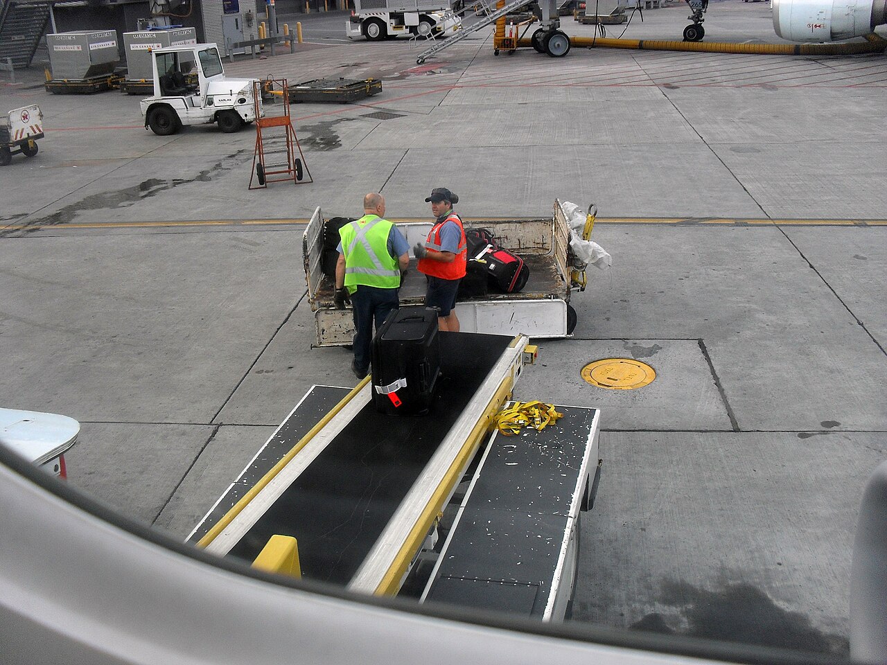 Ramp agents loading suitcases from a baggage cart onto a belt loader beside an airliner