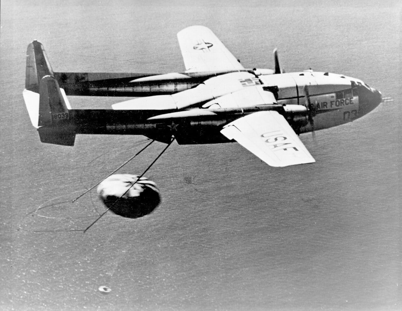 A US Air Force Fairchild C-119J Flying Boxcar snagging a parachute-borne capsule in mid-air with gear trailing from its rear door