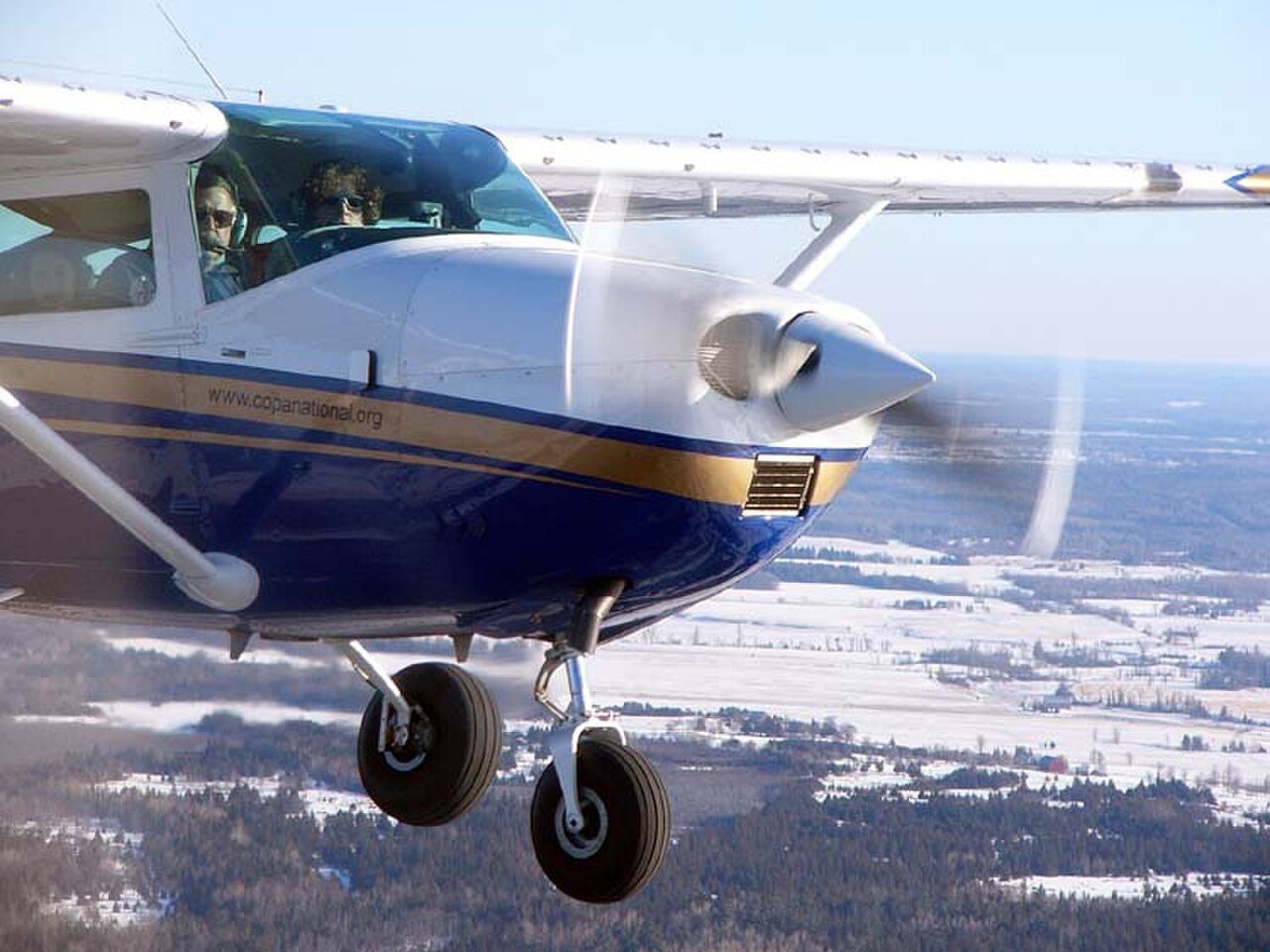 A blue and white Cessna 182 in flight over snowy fields with a row of small vortex generators along the top of its wing