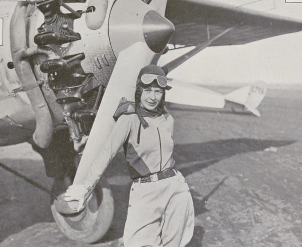 Elinor Smith in flying helmet, goggles and breeches leaning on the propeller of a radial-engined aircraft