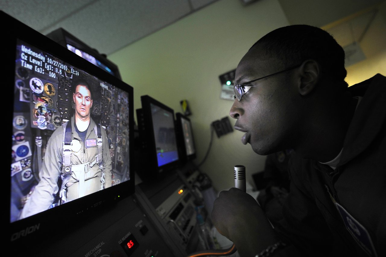 Technician with a microphone coaching an F-16 pilot shown on a centrifuge monitor, with the Gz level readout at 8.6