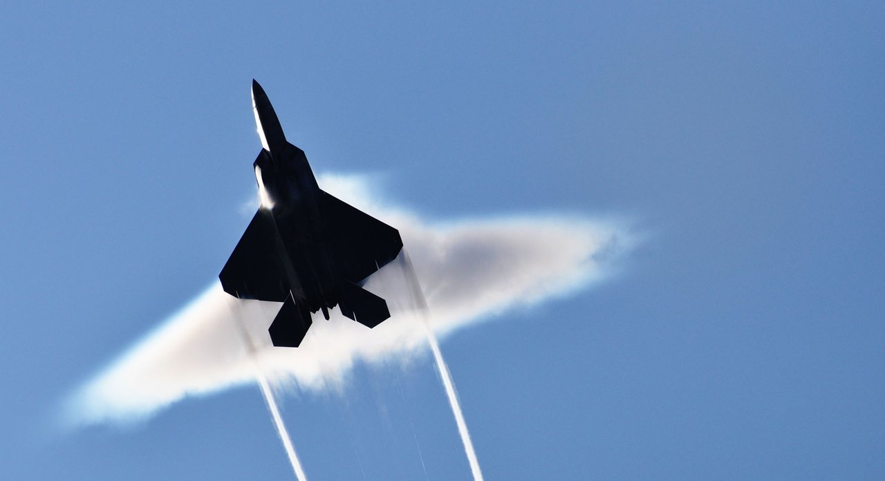 F-22 Raptor seen from below inside a flat disc of vapor, trailing two white vortex lines