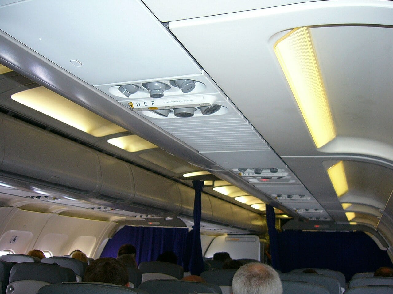 Passengers in an Airbus A320 economy cabin with overhead air outlets and lighting