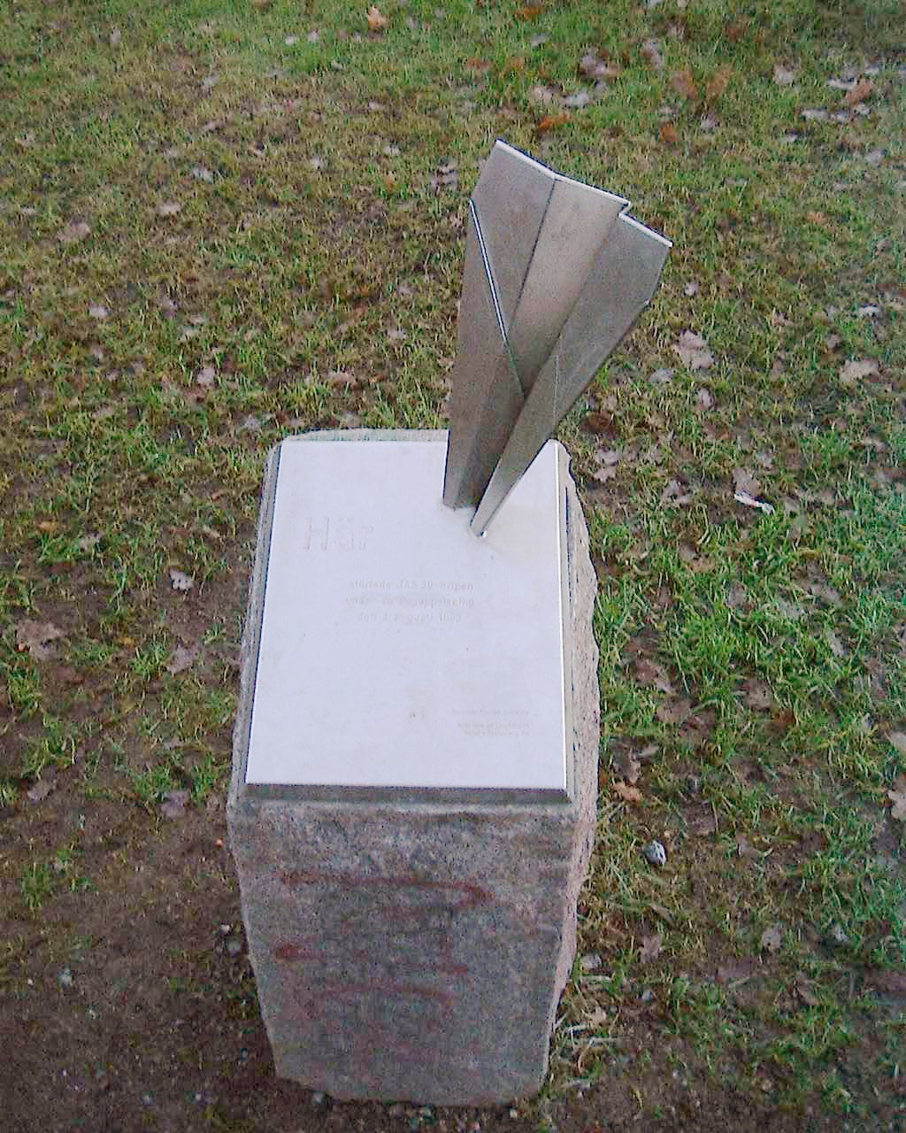 A stainless steel paper-plane sculpture on a stone block on Långholmen, Stockholm, marking the 1993 Gripen crash site