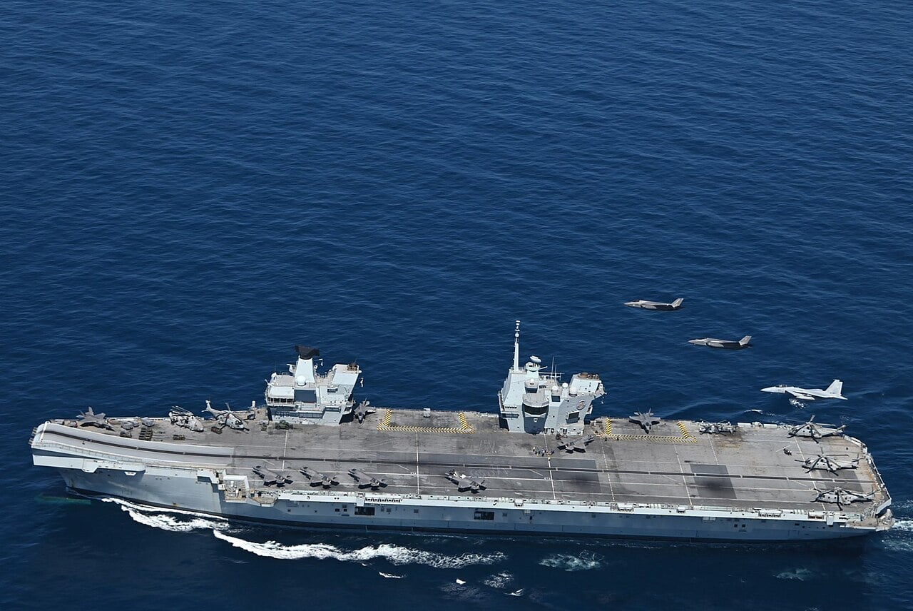 Aerial view of HMS Prince of Wales at sea with F-35B jets and helicopters on deck as fighters fly past