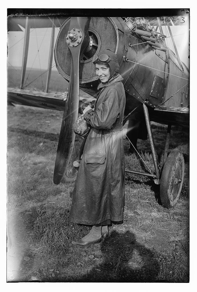 Katherine Stinson in a long leather flying coat, helmet and goggles standing beside the propeller of a biplane