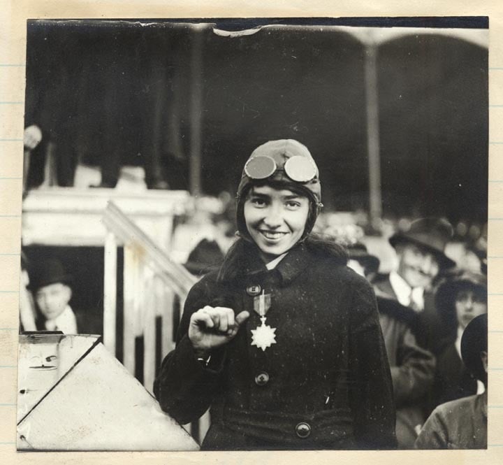 Katherine Stinson in flying helmet and goggles smiling and pointing to a star-shaped medal on her coat at Tanforan race track