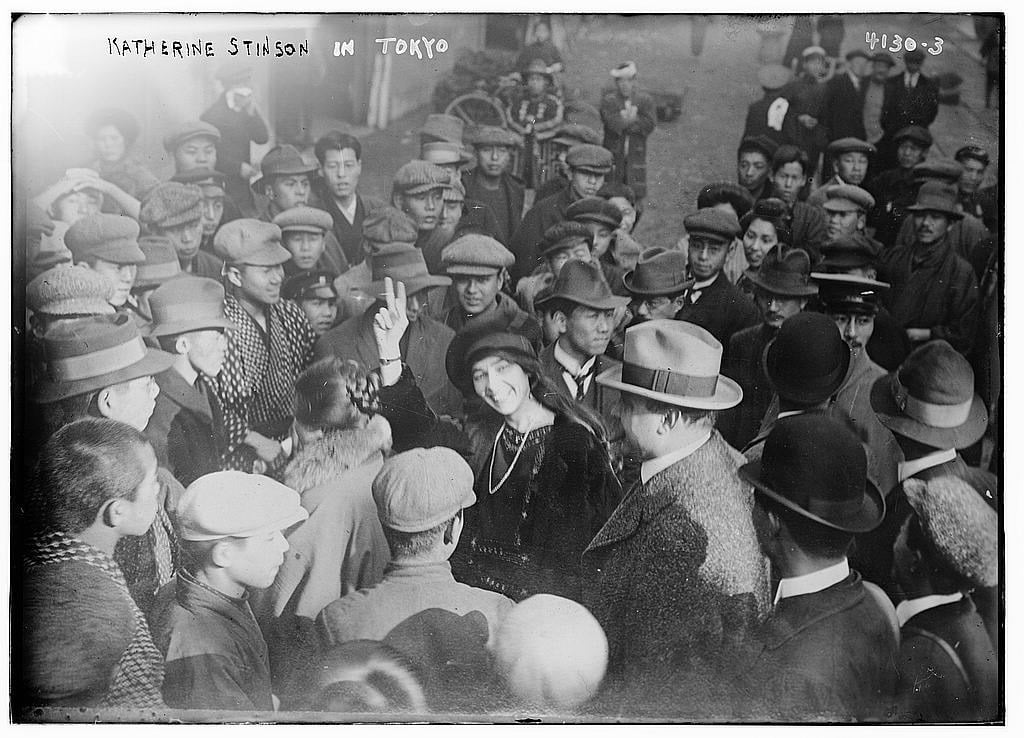 Katherine Stinson smiling and making a V sign in the middle of a large crowd of men in Tokyo
