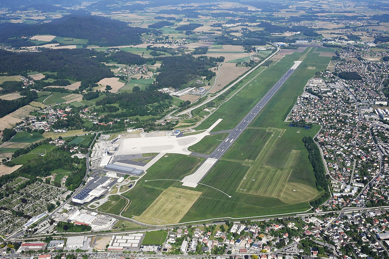 Aerial view of Klagenfurt Airport with its single runway, terminal and apron surrounded by fields and houses