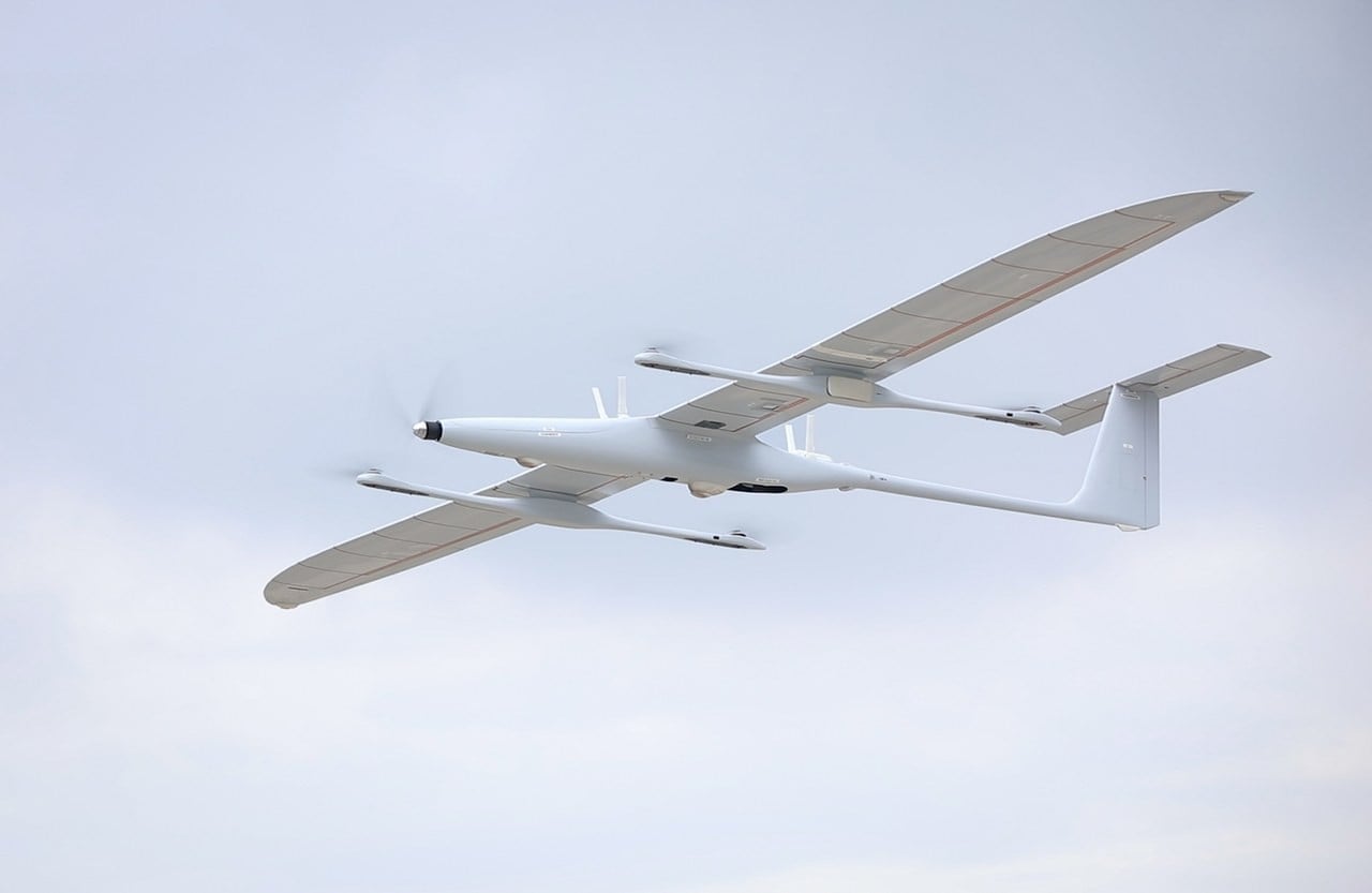 A white Kraus Hamdani K1000 long-endurance drone with a long slender wing in flight against a pale sky