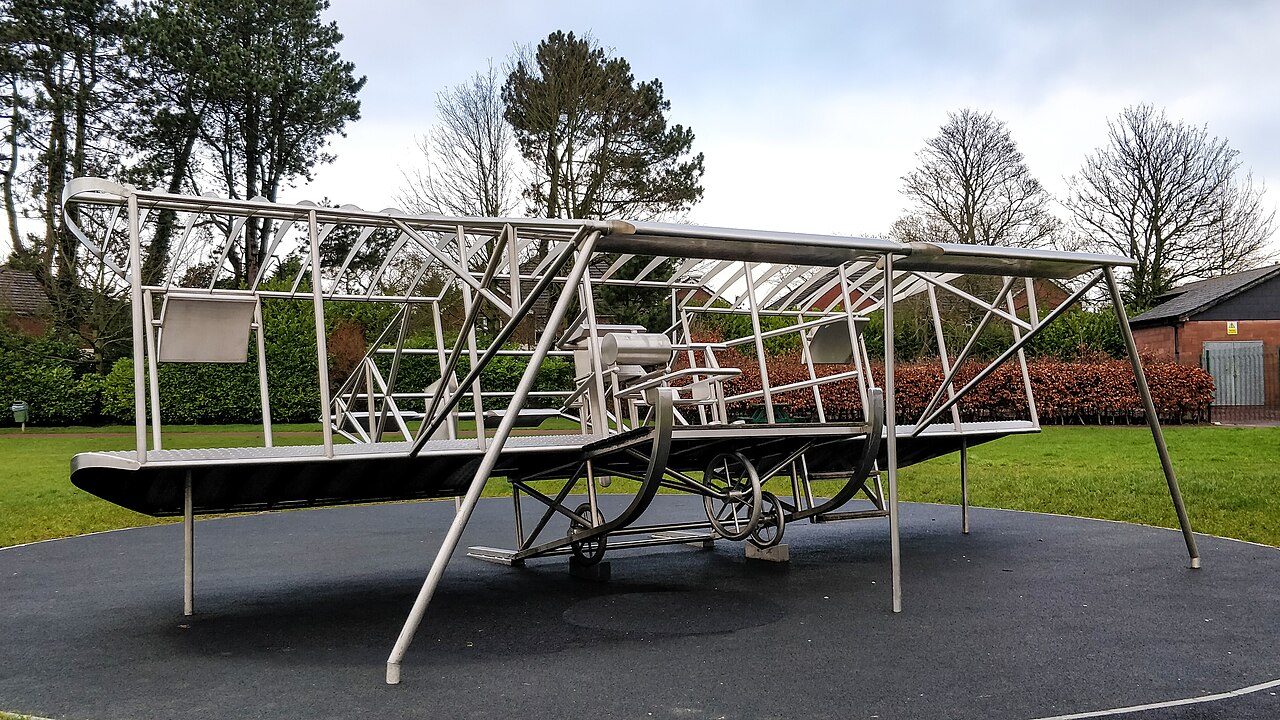 Stainless-steel sculpture of the Mayfly biplane on a round platform in a green park with trees behind