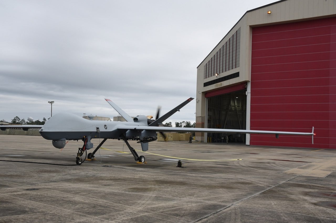A grey MQ-9 Reaper parked on a concrete ramp in front of a red hangar door