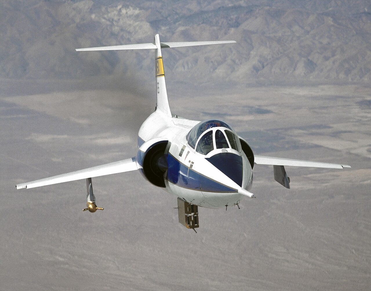 A white NASA F-104 Starfighter in flight over the desert, its short wings angled downward beneath a tall T-tail