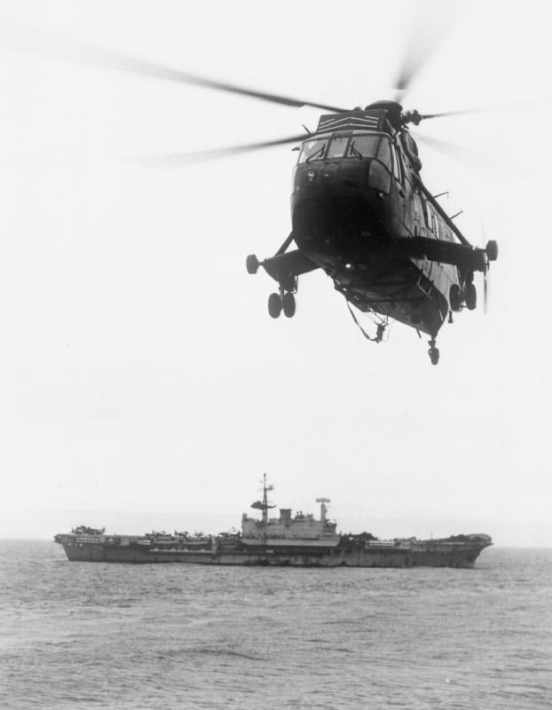 A Royal Navy Westland Sea King helicopter hovering with the aircraft carrier HMS Hermes in the background