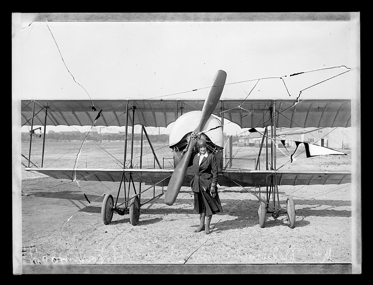 Raymonde de Laroche in a long coat and flying helmet standing in front of the propeller of a Farman biplane on an airfield in 1919