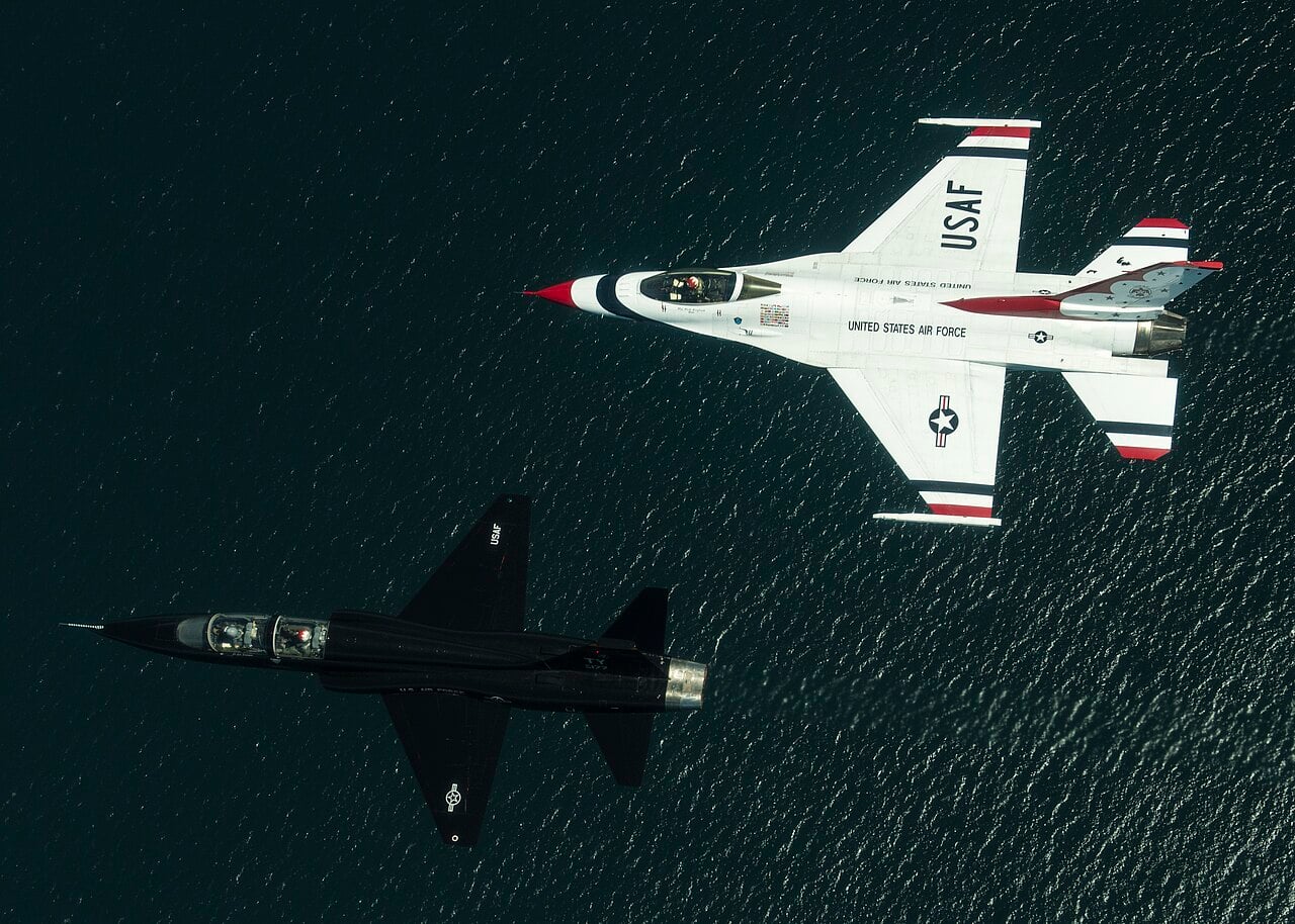 A Thunderbirds F-16 and a black T-38 Talon flying a two-ship formation over the water near Tyndall Air Force Base