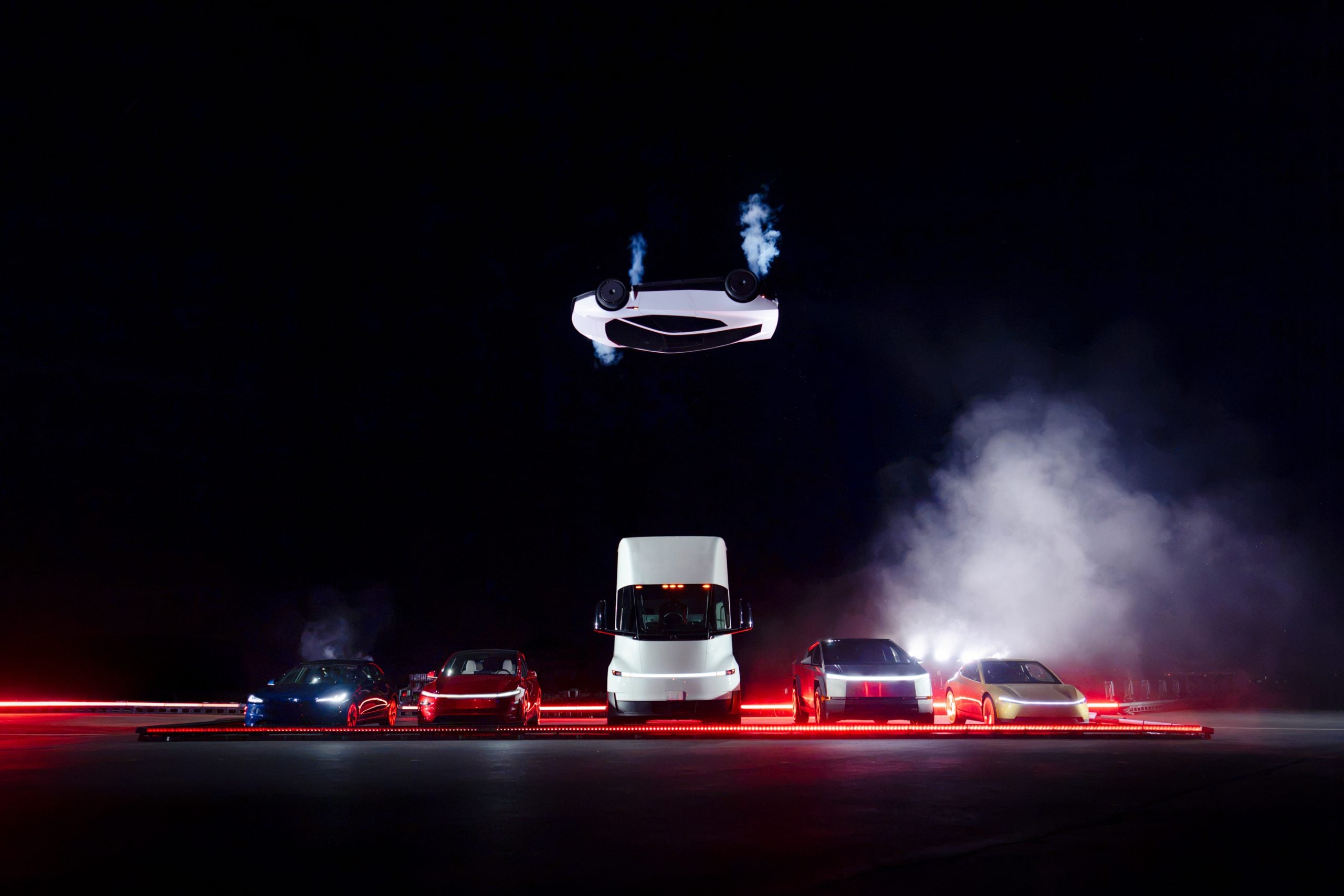 The new Tesla Roadster inverted in the air above a line of Tesla vehicles at McGregor, Texas, with thruster plumes visible